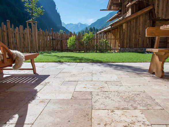 Sol de la terrasse en travertin rustique et meubles en bois, au fond une pelouse et vue sur les montagnes