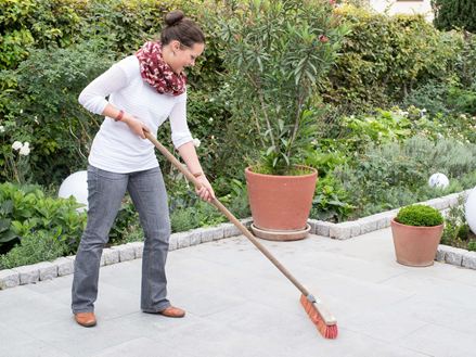 Une femme nettoie une terrasse en granit avec un balai, en arrière-plan un jardin naturel